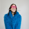 Susie wearing a blue fur jacket and pink sunglasses. She is grinning while holding a silver magic wand with her teeth.