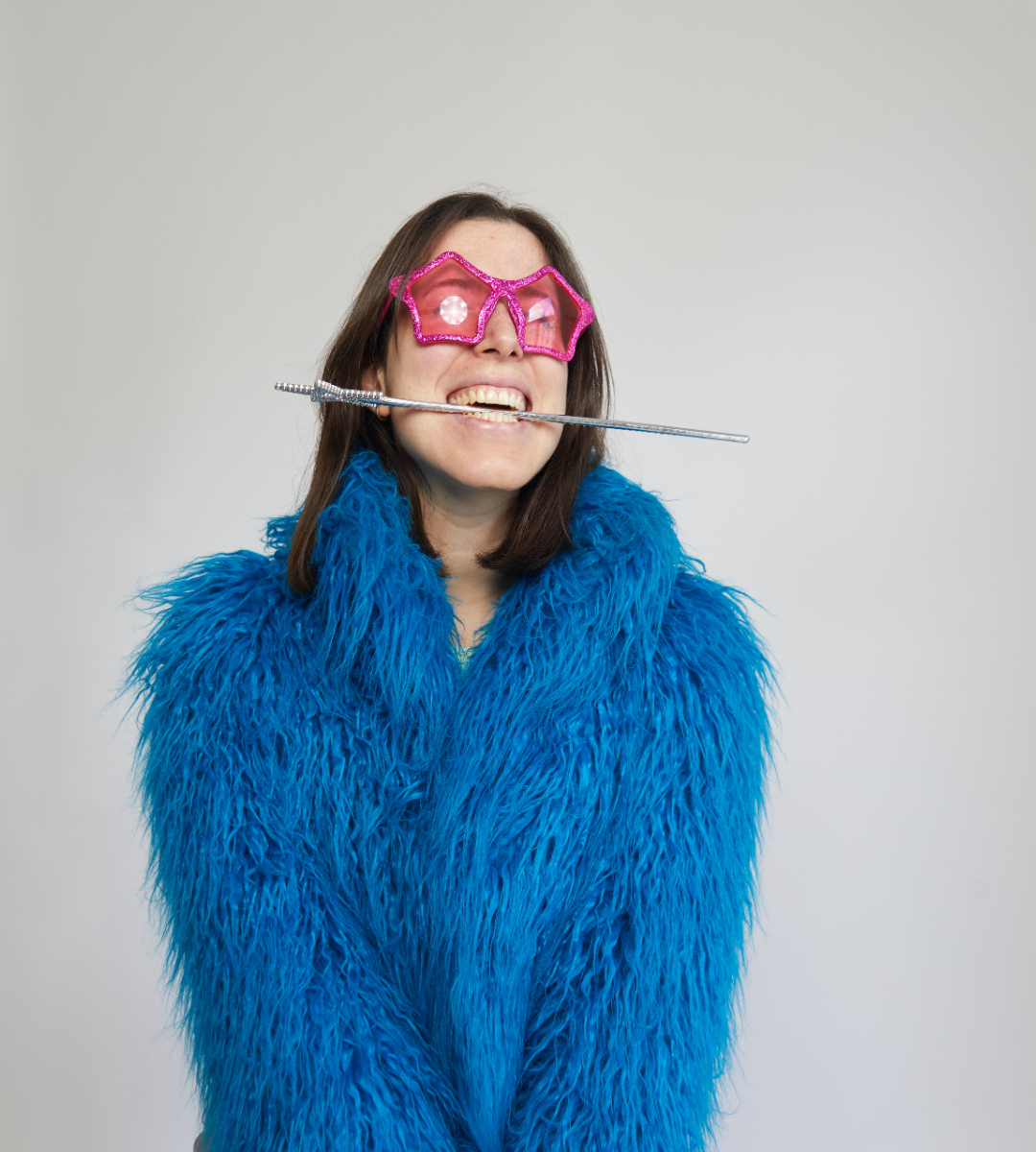 Susie wearing a blue fur jacket and pink sunglasses. She is grinning while holding a silver magic wand with her teeth.