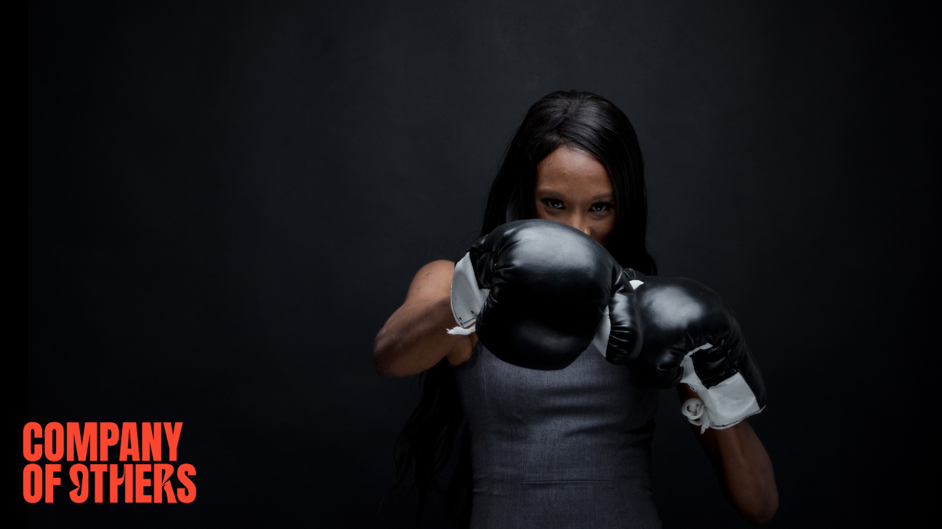 A young woman in boxing gloves punches towards the camera playfully against a dark background. Company of Others logo sits in the bottom left corner.