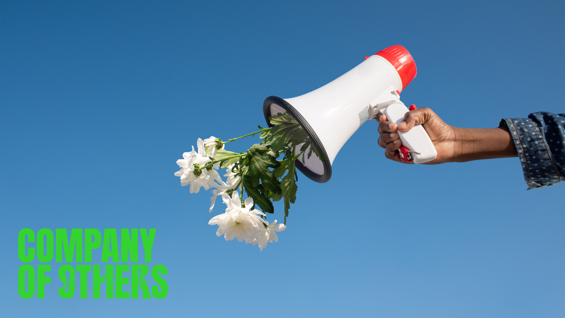 A hand holds a megaphone with flowers coming out of it against the sky. The Company of Others logo sits in the bottom left-hand corner.